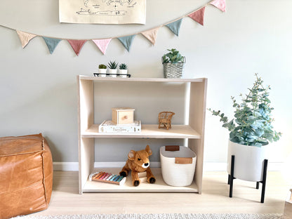 A wooden toy shelf with three tiers, displaying various toys and decorative items like a potted plant and a teddy bear, in a room with a leather ottoman, a white wall, and decorative flags hanging from the ceiling.