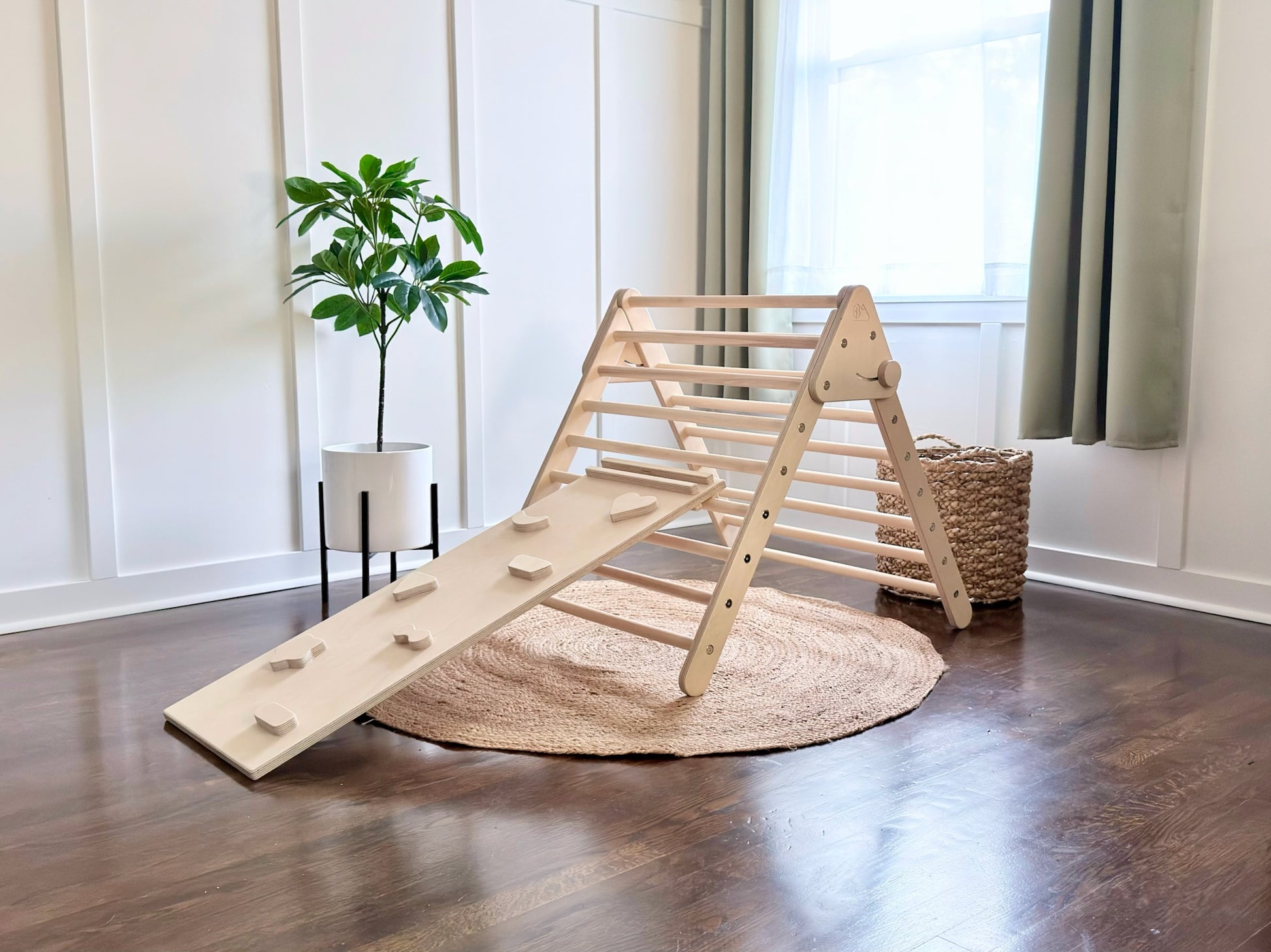 A natural wood climbing frame with an attached ramp and small pegs, set up on a round jute rug in a bright room with hardwood floors, a potted plant, and a window.