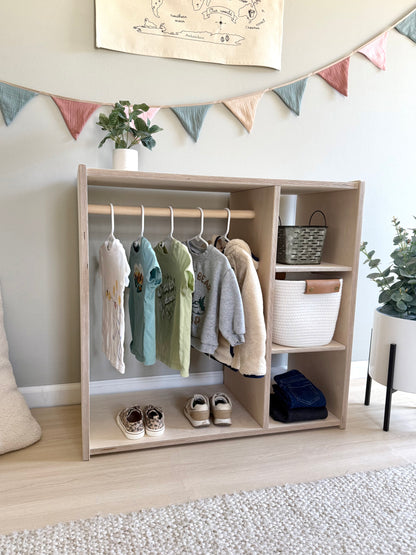 Children's Montessori style wardrobe with clothes hanging on the bar, shoe rack, and storage compartments, placed in a room with decorative banners on the wall.
