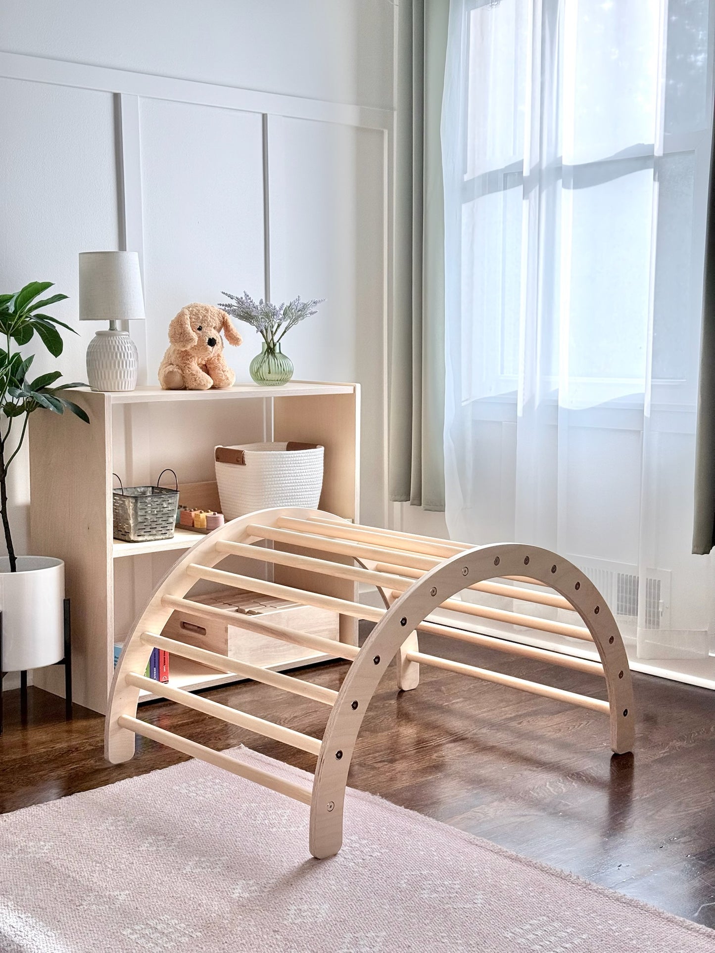 A light natural wood arched climbing or play structure with curved legs and slatted support bars, shown in a bright child's room with a rug, shelving, window, and decor.