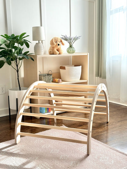 A light natural wood arched play gym or climbing frame with curved bars, shown in a bright, modern nursery or playroom setting with a wooden floor, rug, shelving, and plants.