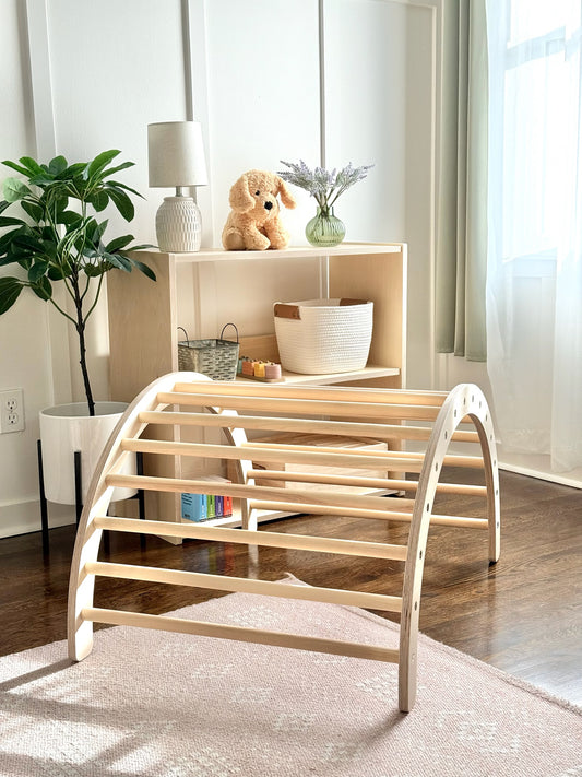 A light natural wood arched play gym or climbing frame with curved bars, shown in a bright, modern nursery or playroom setting with a wooden floor, rug, shelving, and plants.