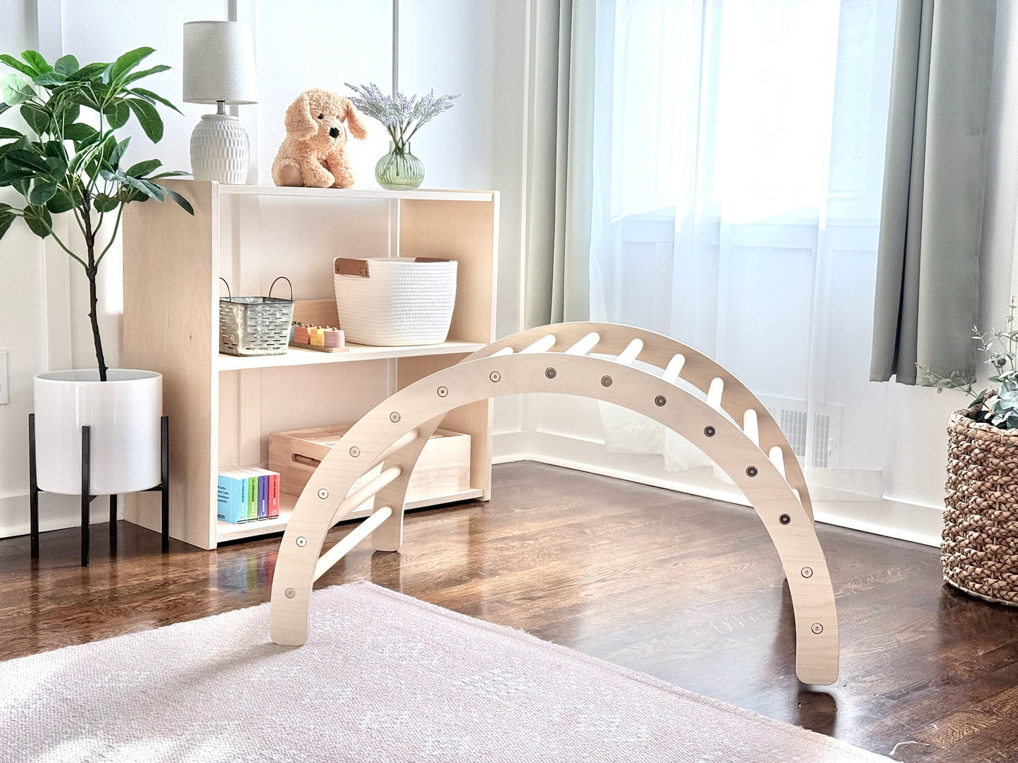 A light natural wood arched play structure with circular cutouts stands on a pale rug in a bright, modern room with a wooden floor, white shelving, and plants.