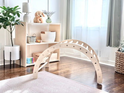 A light natural wood arched play structure with circular cutouts stands on a pale rug in a bright, modern room with a wooden floor, white shelving, and plants.