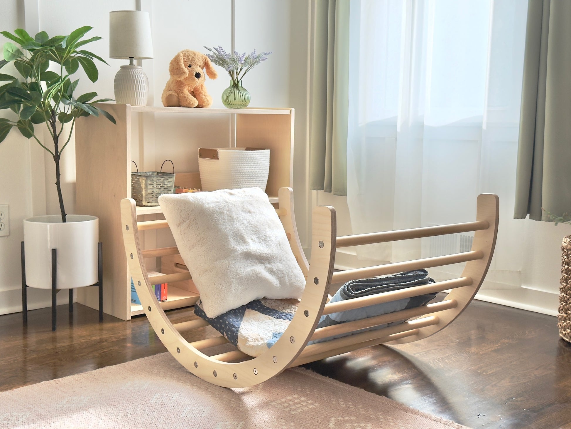 A light natural wood rocking cradle with a curved backrest and front safety slats, styled with a white throw pillow and folded blankets in a bright nursery room.
