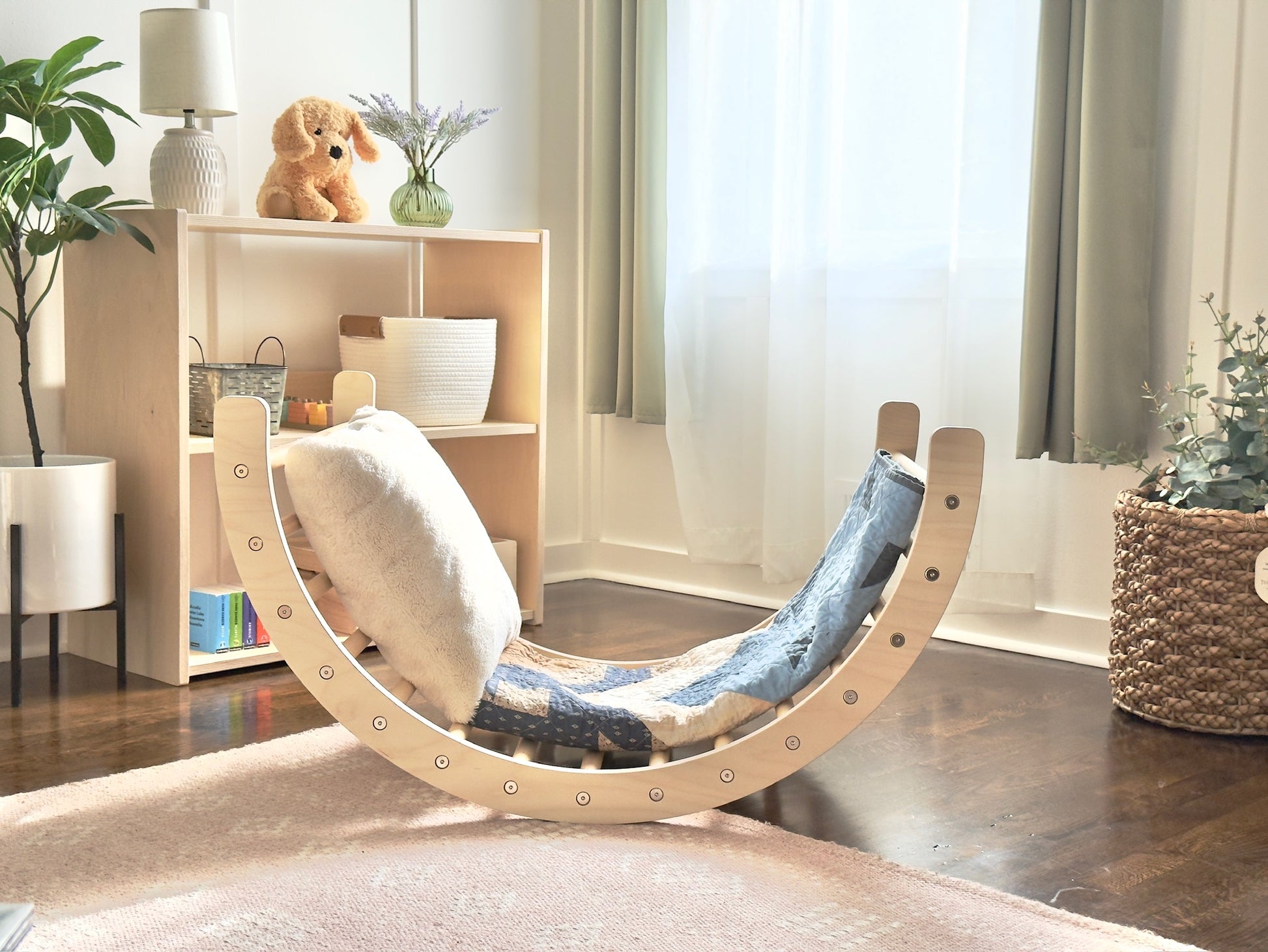 A crescent-shaped wooden rocking chair with a blue and white patterned fabric seat, styled in a bright, modern nursery or playroom with wooden floors, a light wood shelf, and sheer curtains.