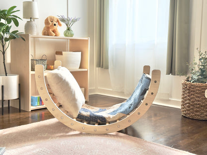 A crescent-shaped wooden rocking chair with a blue and white patterned fabric seat, styled in a bright, modern nursery or playroom with wooden floors, a light wood shelf, and sheer curtains.