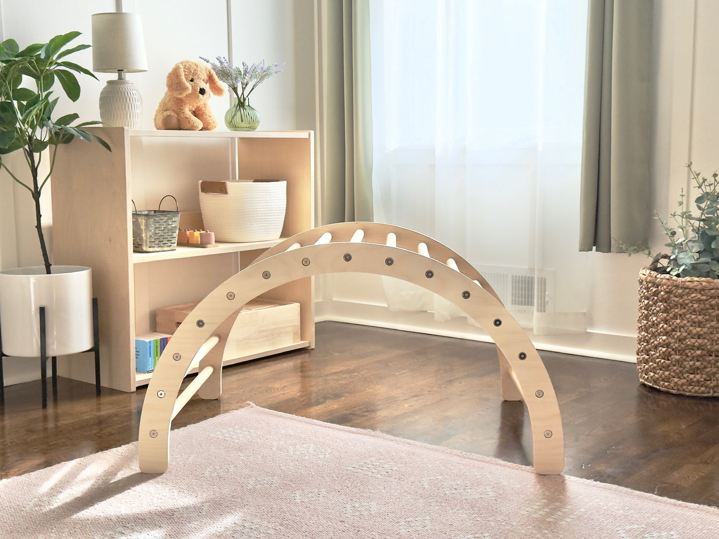 A light natural wood arched play structure with circular cutouts, displayed on a pale rug in a bright, modern nursery or playroom setting.