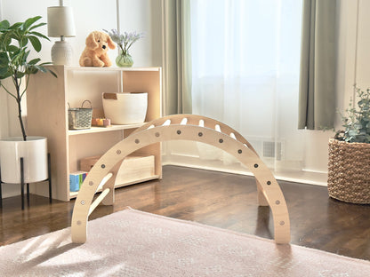 A light natural wood arched play structure with circular cutouts, displayed on a pale rug in a bright, modern nursery or playroom setting.
