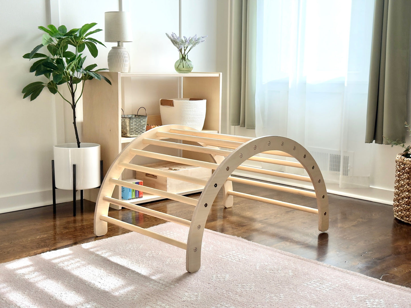 A light natural wood arched play gym or climbing frame with curved support bars, shown in a bright, modern room with a rug, shelving, and plants.