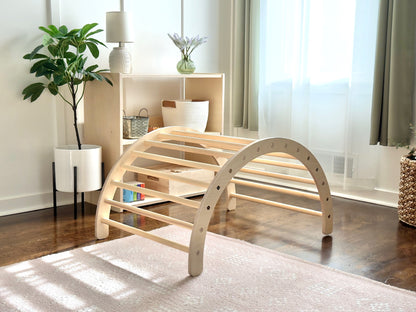 A light natural wood arched play gym or climbing frame with curved support bars, shown in a bright, modern room with a rug, shelving, and plants.