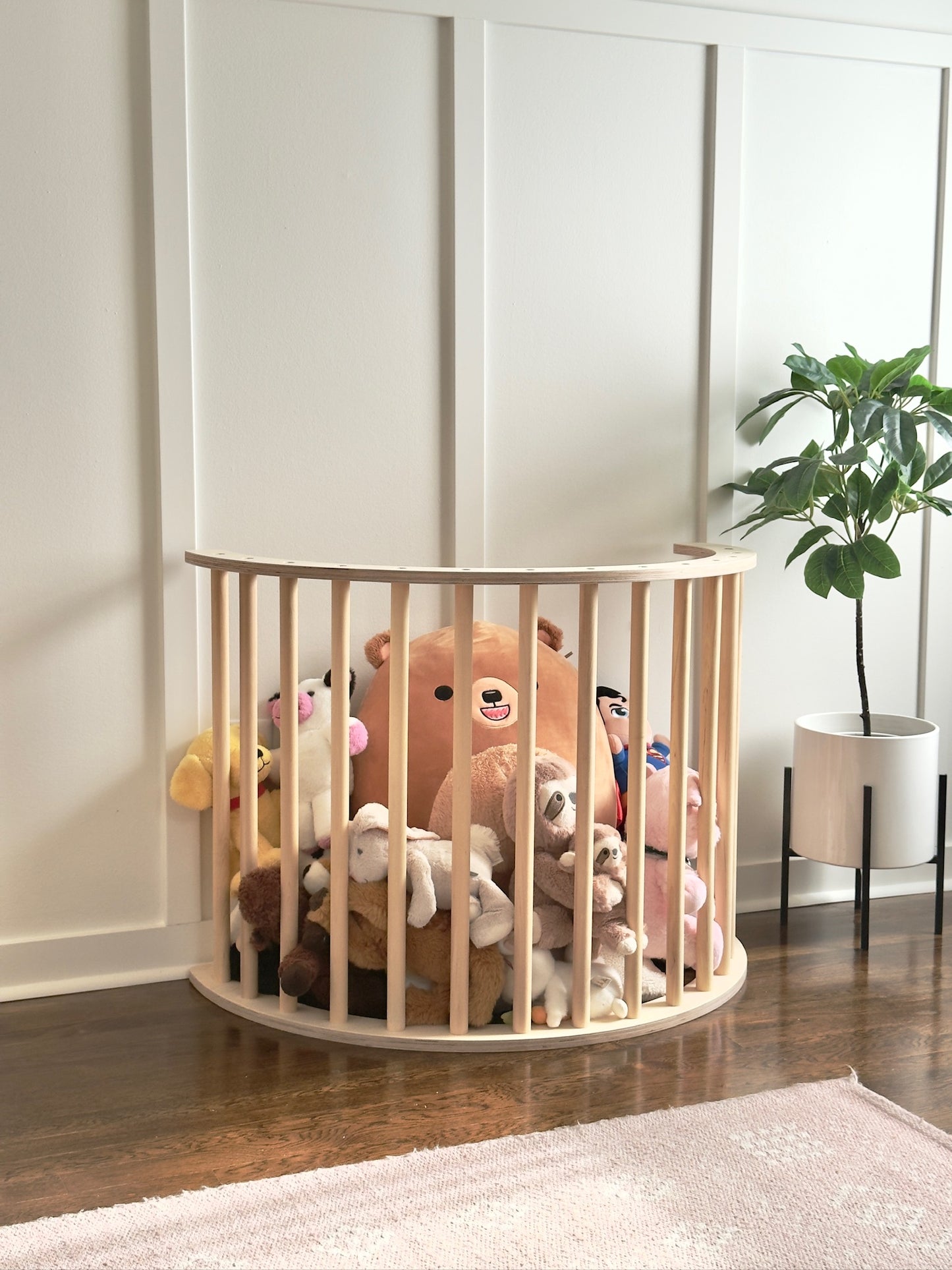 A light natural wood oval crib or playpen with vertical slats, filled with assorted plush stuffed toys, set on a dark wood floor against a white paneled wall with a potted plant nearby.