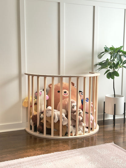 A light natural wood oval crib or playpen with vertical slats, filled with assorted plush stuffed toys, set on a dark wood floor against a white paneled wall with a potted plant nearby.
