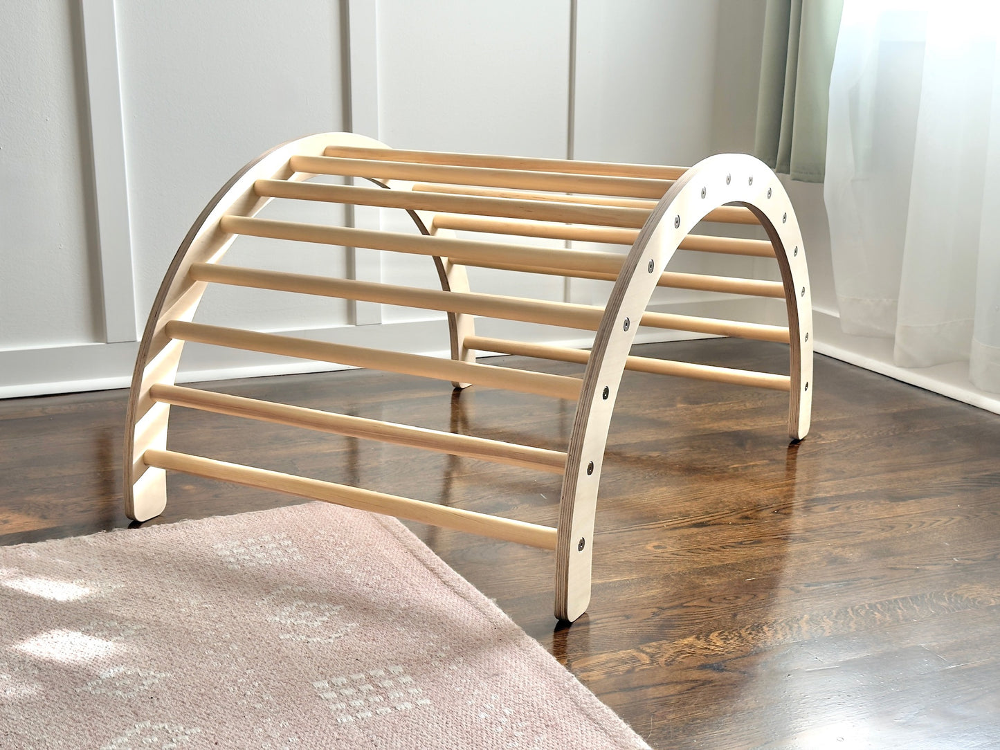 A light natural wood arch-shaped pet play mat with horizontal slats, shown on a dark wood floor in a room setting.