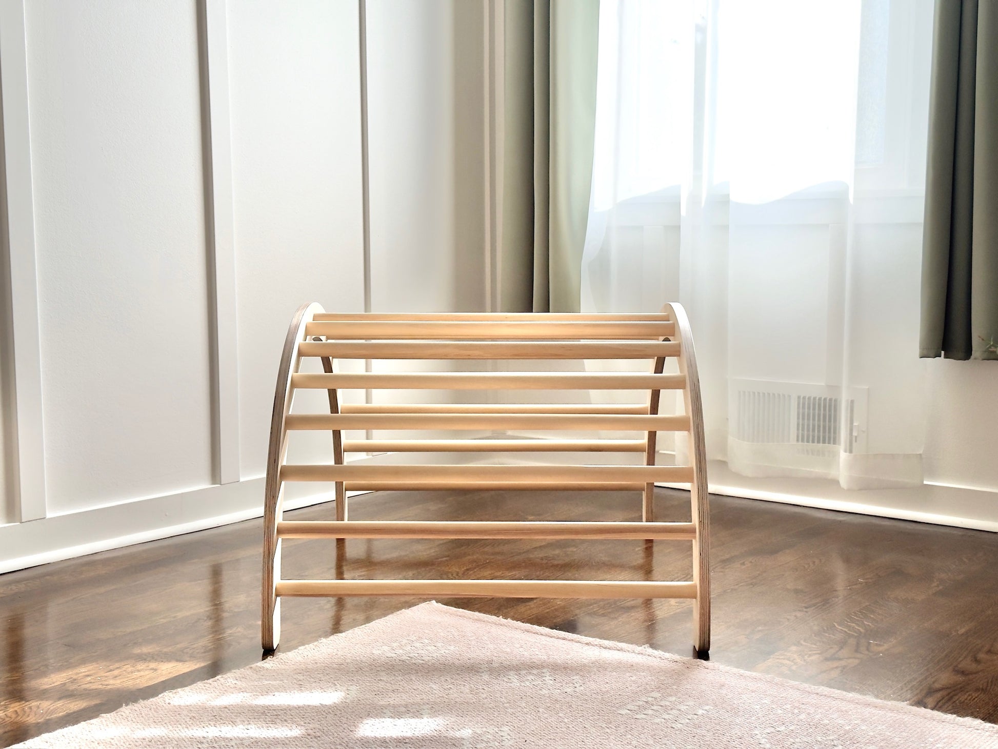 A light natural wood montessori climbng toy with curved side supports and multiple horizontal slats, positioned on a dark wood floor in a bright room with white paneled walls and sheer curtains.