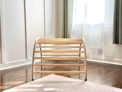 A light natural wood montessori climbng toy with curved side supports and multiple horizontal slats, positioned on a dark wood floor in a bright room with white paneled walls and sheer curtains.