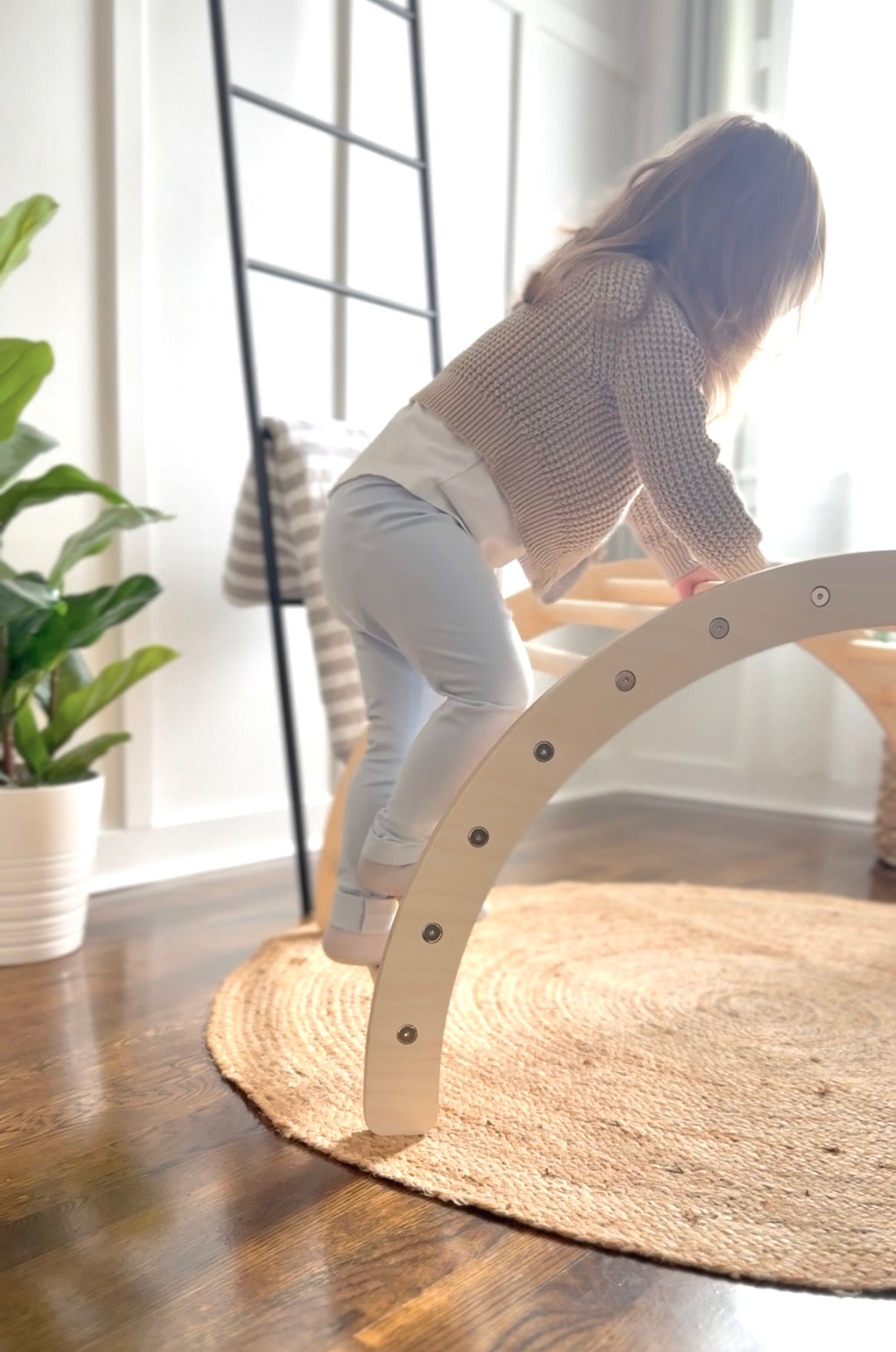 A young child in a beige knit sweater and light blue pants climbs on a curved, light-colored wooden playground structure on a round jute rug over a hardwood floor.