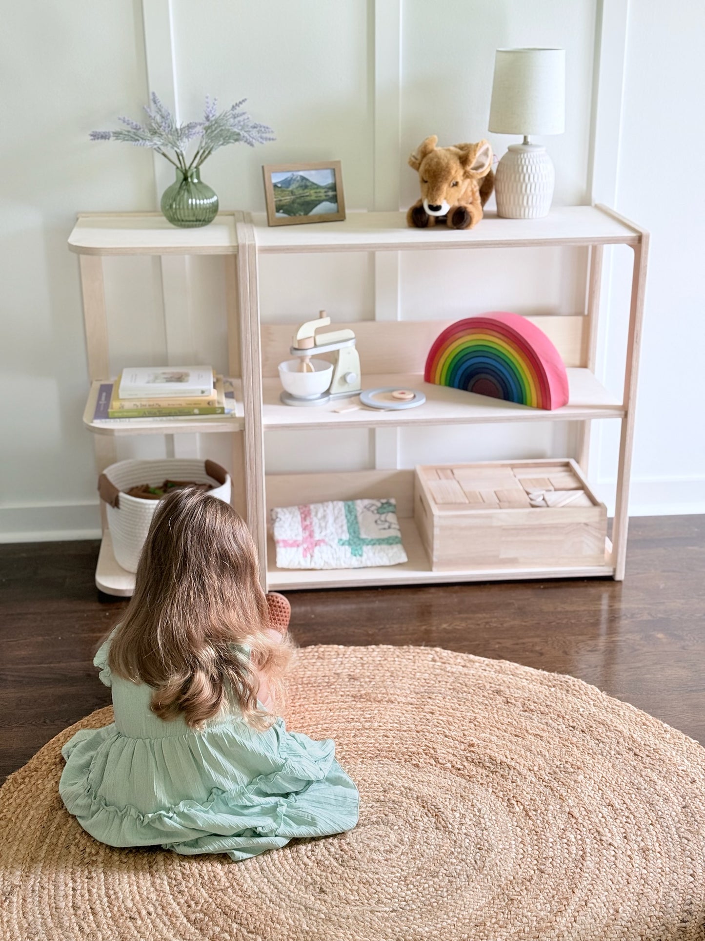 A young child sits on a round woven rug in front of a light wood Montessori shelving unit filled with toys and books in a bright room.