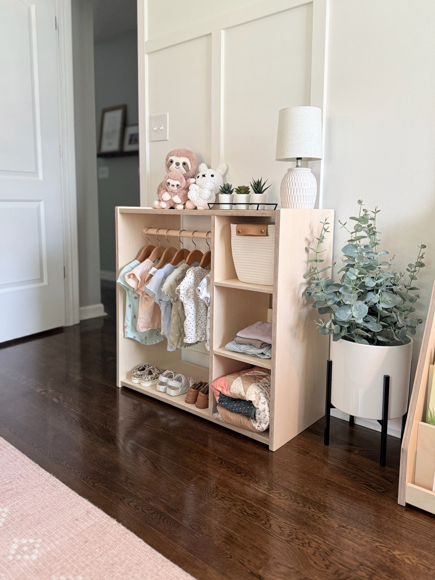 Wooden storage unit with baby clothes and shoes in a room with a lamp and plant.