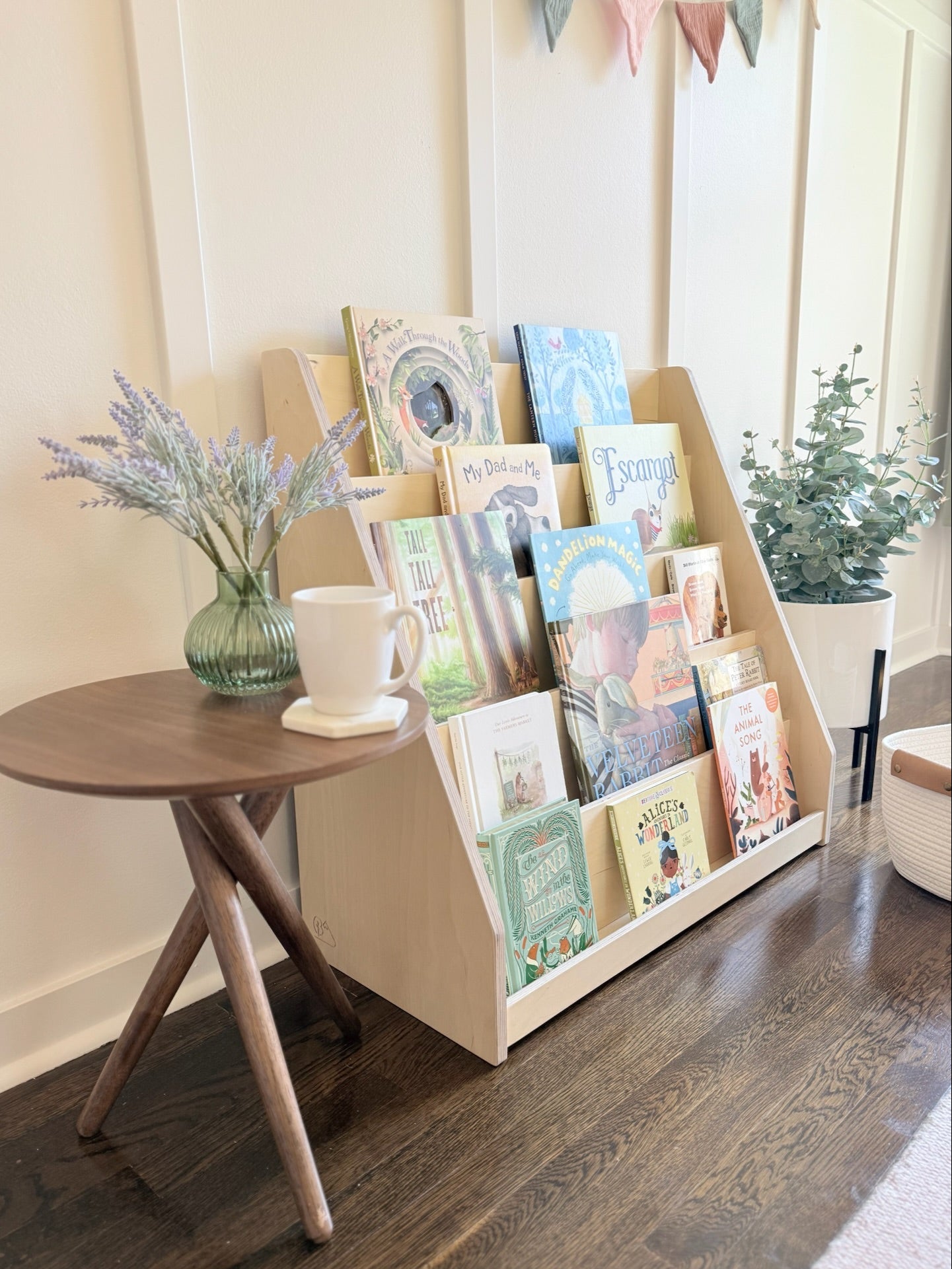 Children's books displayed in a wooden bookshelf next to a small round table with a mug and vase.