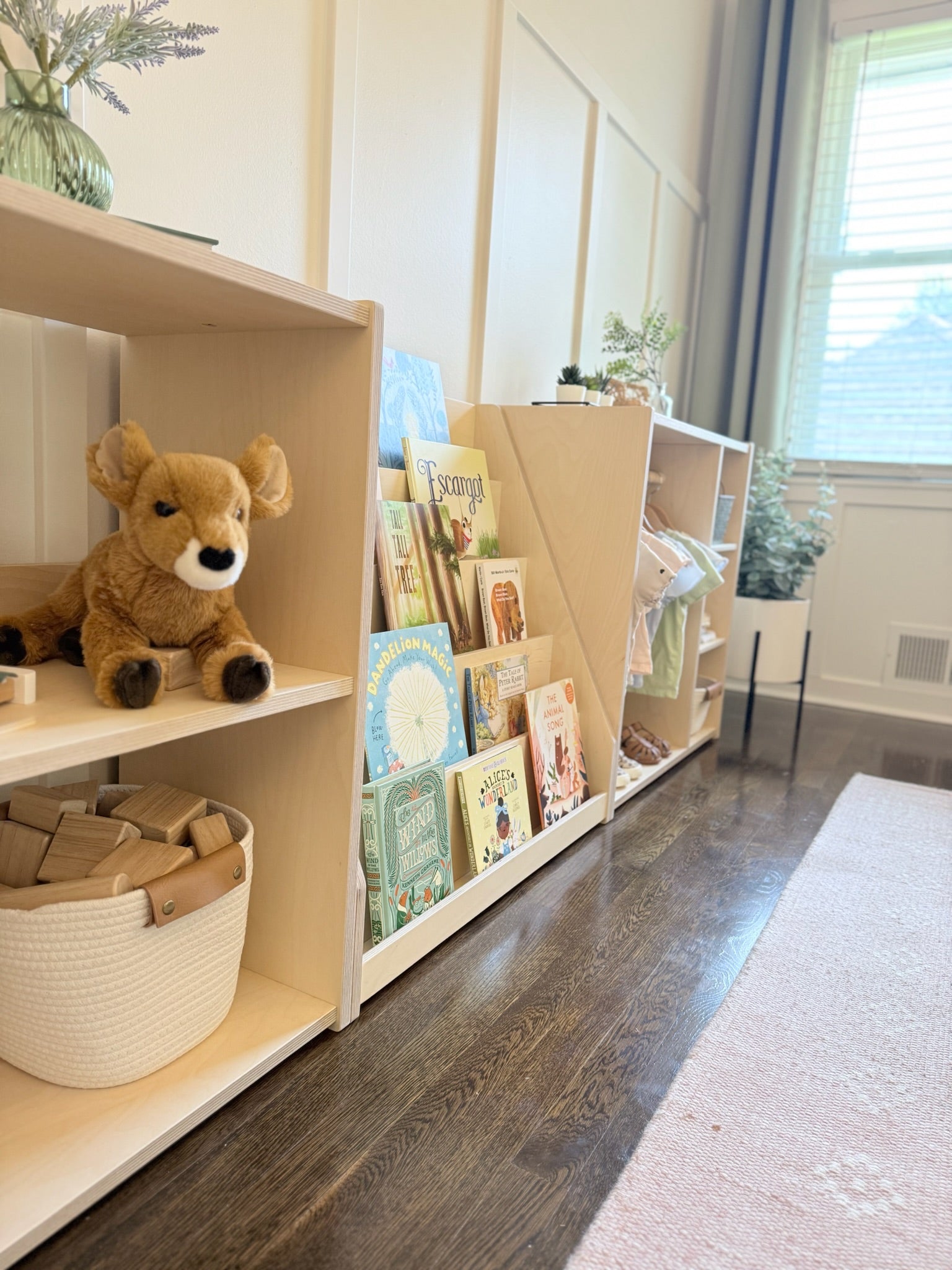 Children's bookshelf with books and a teddy bear in a room with a window and rug.
