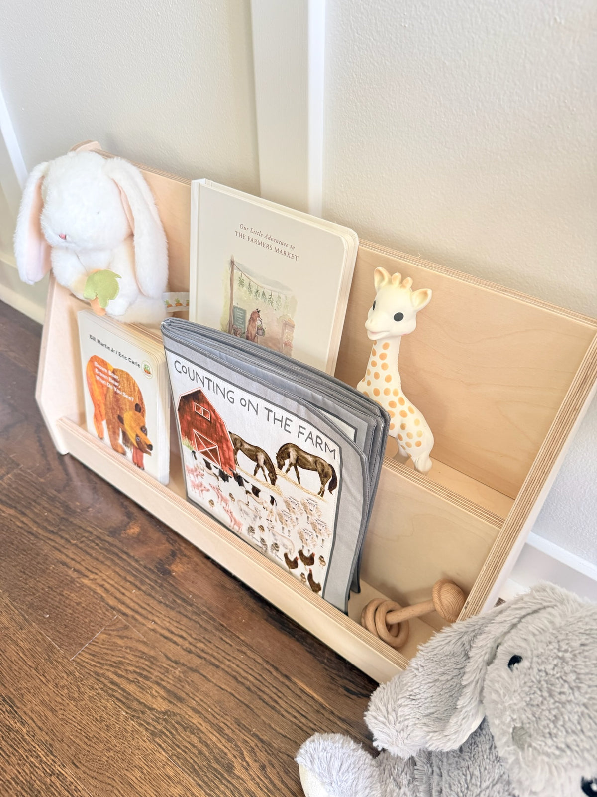 A light natural wood children's bookshelf organizer on a dark wood floor, styled with a white plush bunny, a giraffe toy, a gray book, and other children's books.