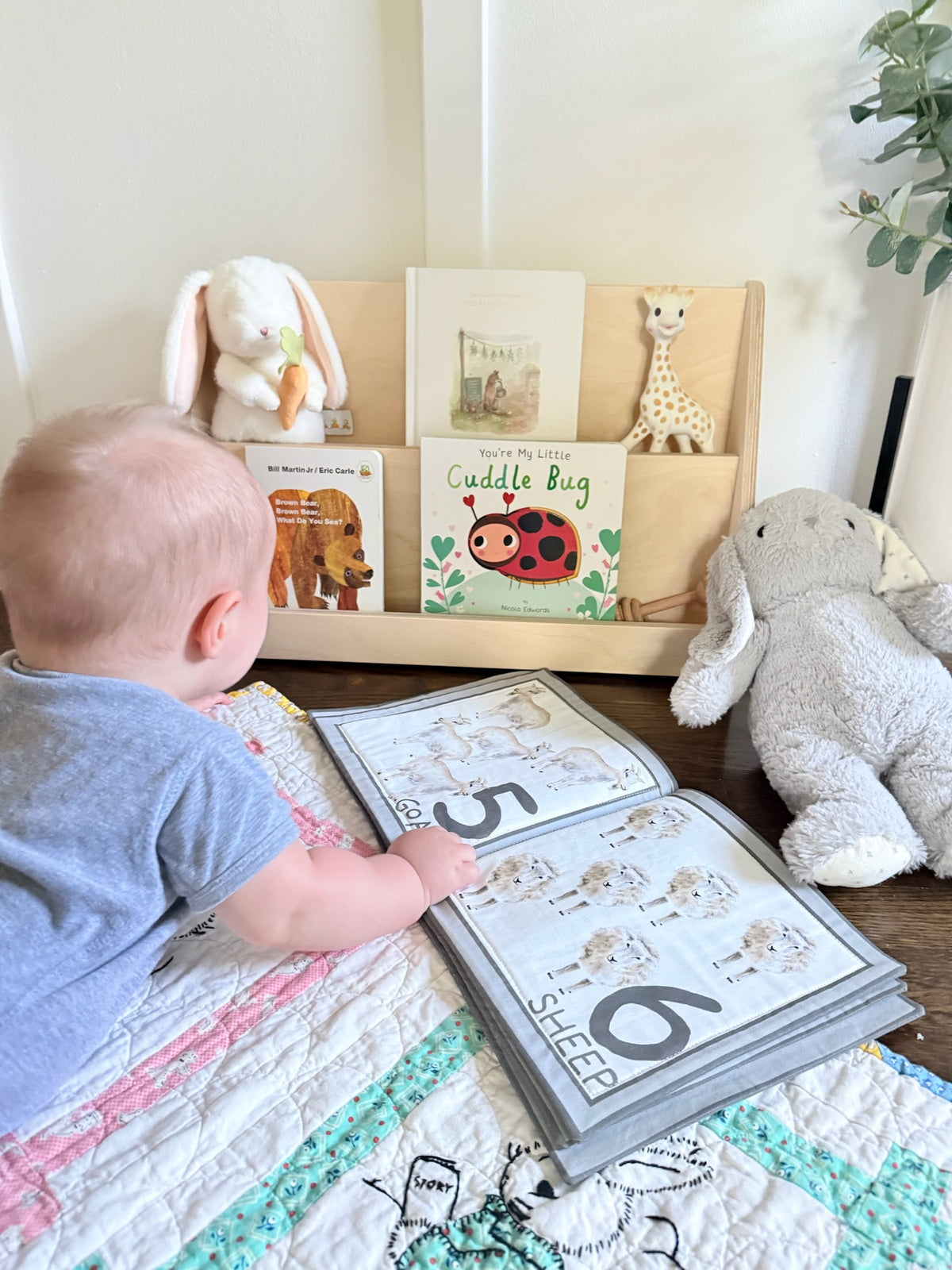 A baby in a blue shirt sits on a quilted blanket, looking at an open alphabet learning book. In the background, a wooden bookshelf holds children's books, a plush bunny, a giraffe toy, and a large gray stuffed elephant.