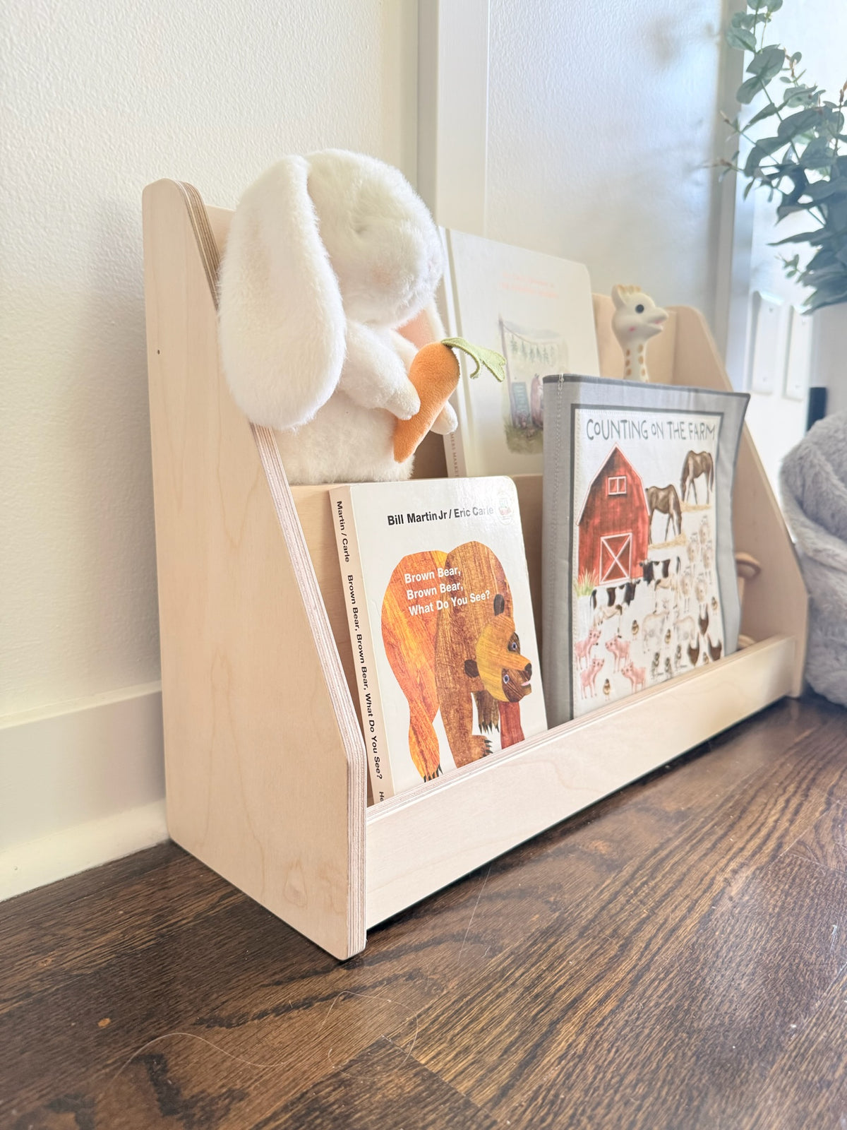 A light natural wood children's bookshelf with three open compartments, styled with a white plush bunny holding an orange carrot and children's books on a dark wood floor against a white wall.