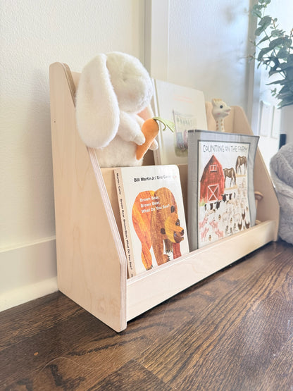 A light natural wood children's bookshelf with three open compartments, styled with a white plush bunny holding an orange carrot and children's books on a dark wood floor against a white wall.