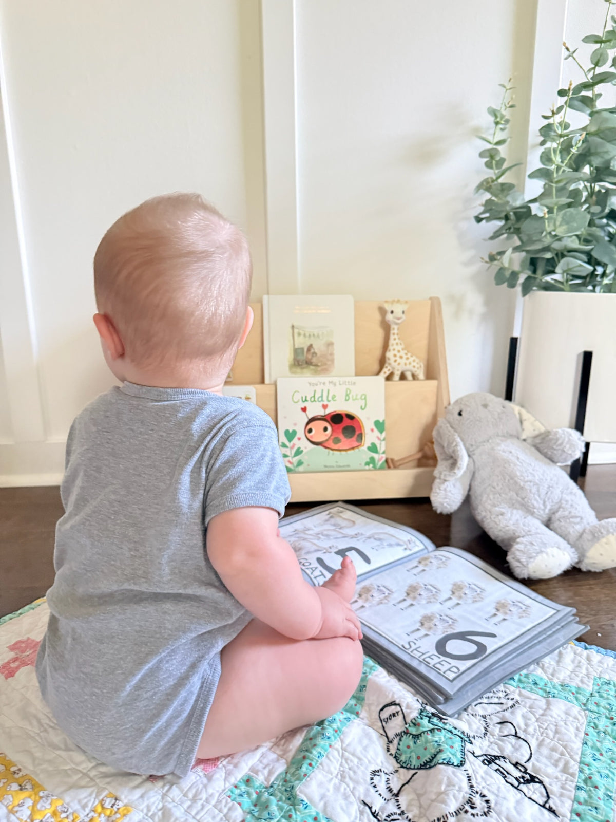 A baby sitting on a quilted blanket, viewed from behind, wearing a heather gray short-sleeve bodysuit. In front of the baby is an open children's activity book, with a small wooden bookshelf holding books and toys in the background.