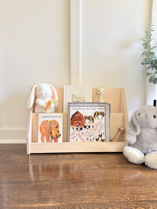 A light natural wood children's book and toy organizer on a dark wood floor, styled with plush toys and children's books in a bright room.