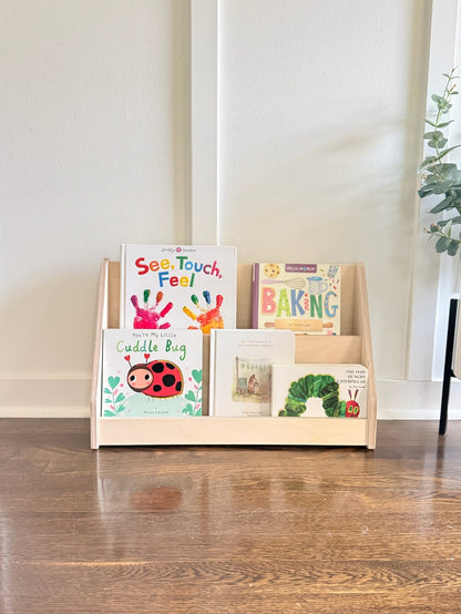 A light natural wood children's book display shelf holding several colorful children's books, placed on a polished wooden floor against a white paneled wall with a potted plant partially visible on the right.