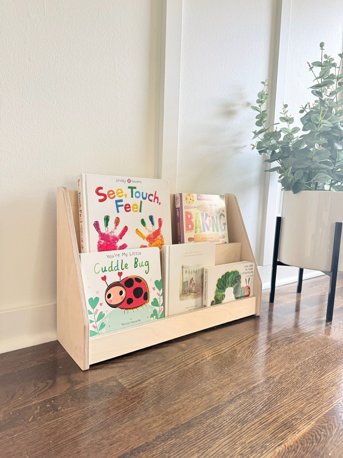 A light natural wood children's book display shelf holding several colorful children's books, placed on a dark wood floor against a white wall with a potted plant in the background.