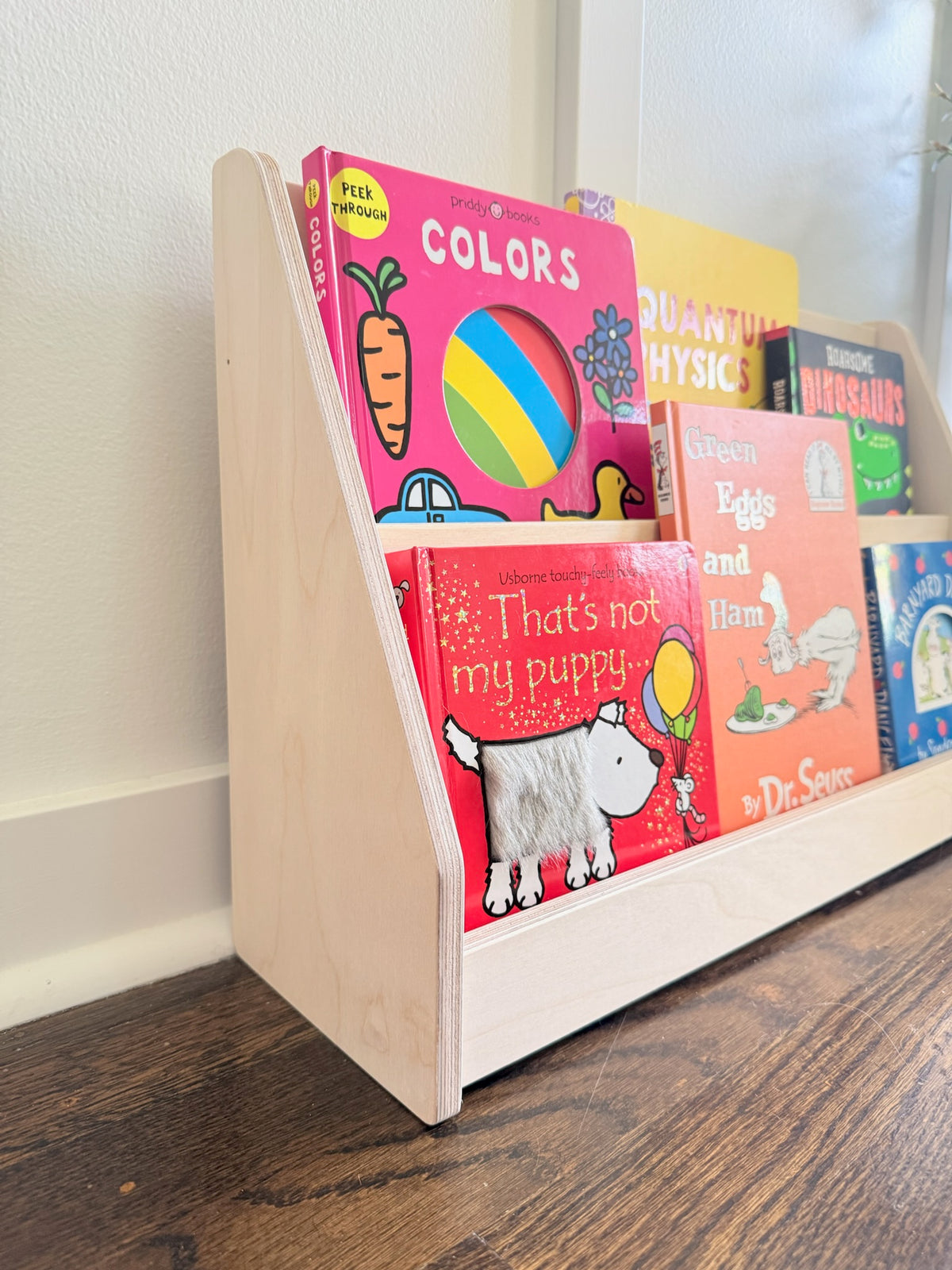 A light natural wood bookshelf on a dark wood floor, holding several colorful children's books against a white wall.