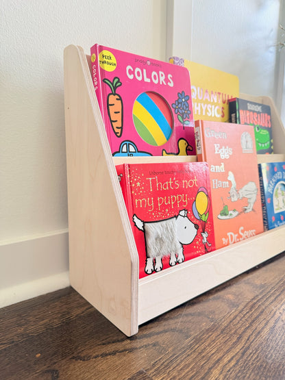 A light natural wood bookshelf on a dark wood floor, holding several colorful children's books against a white wall.
