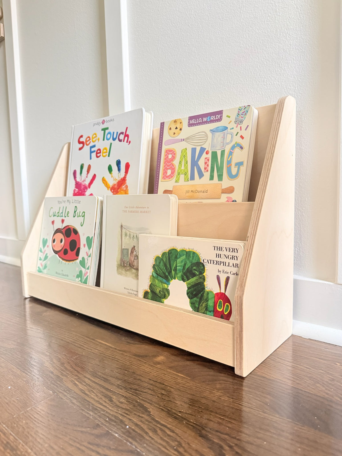 A light natural wood children's book organizer filled with colorful children's books, placed on a dark wood floor against a white wall.
