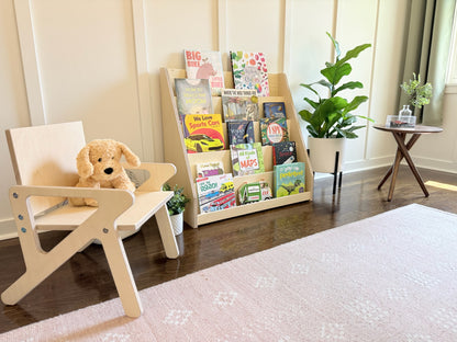 Children's bookshelf with books and a teddy bear on a wooden montessori mid century modern style chair in a room.