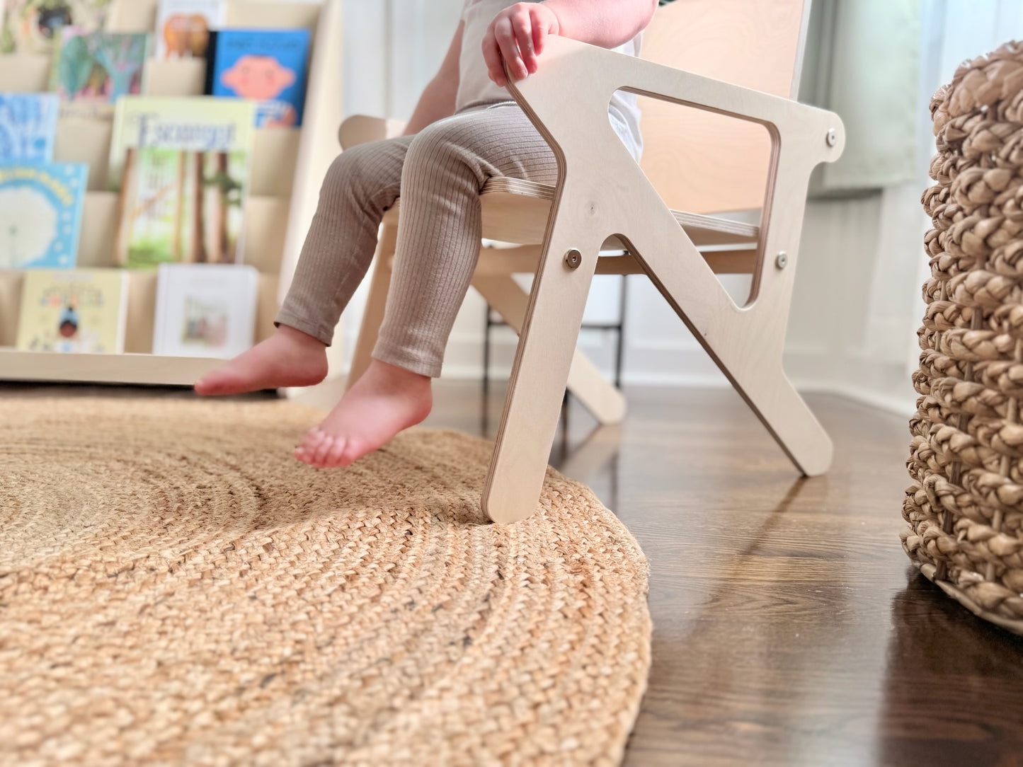 Child sitting on a wooden birch mid century modern style montessori chair in a home setting with books and a rug.