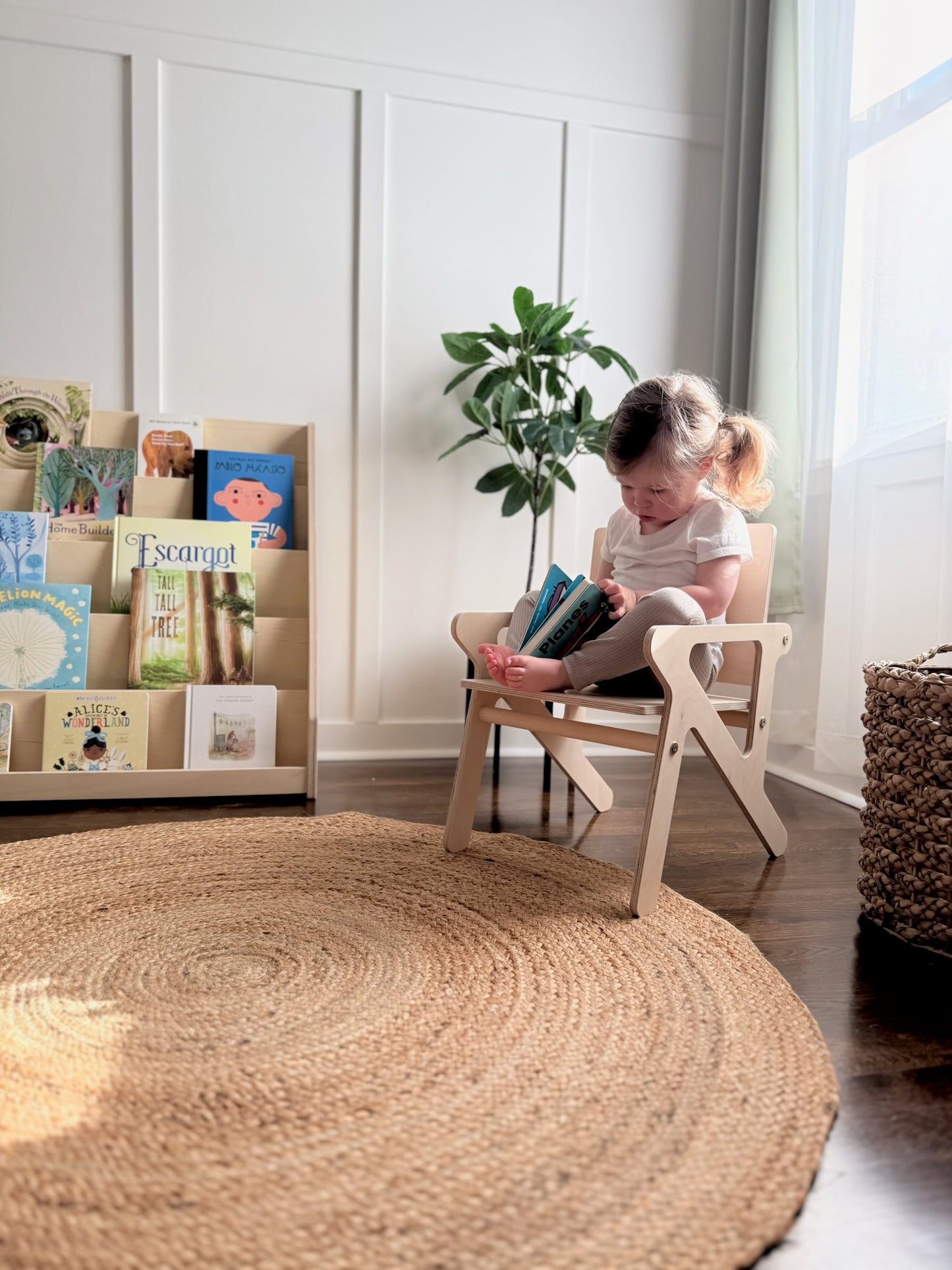 Child reading a book in a wooden montessori mid century modern style chair next to a bookshelf and round woven rug in a bright room.