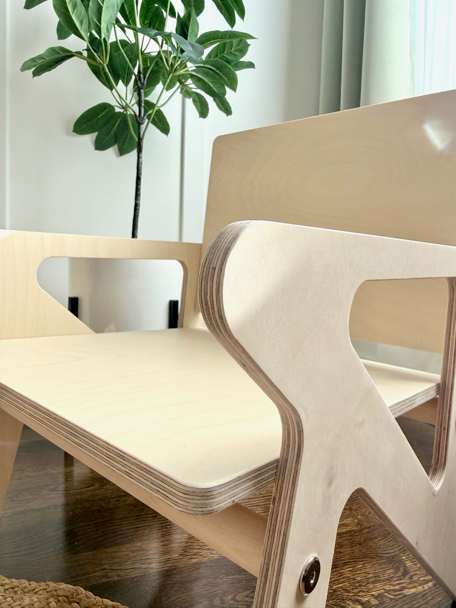 Wooden toddler chair with a modern design in a room with a plant and curtains.