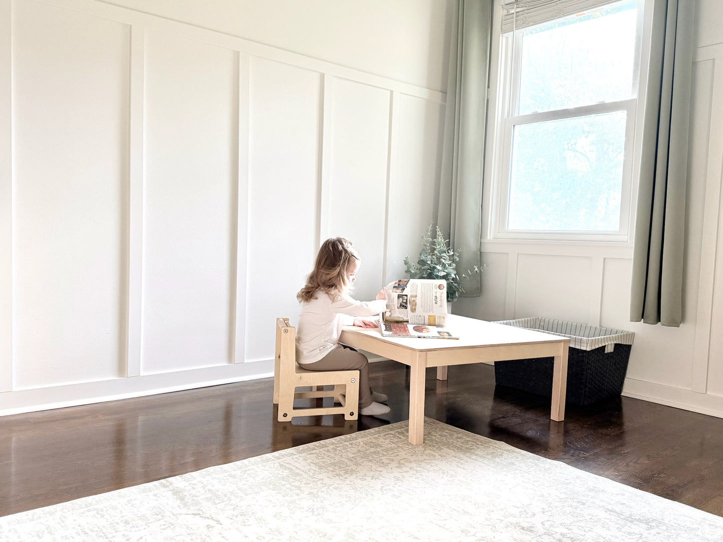 Toddler sitting at a small low wooden table in a bright room with large windows.