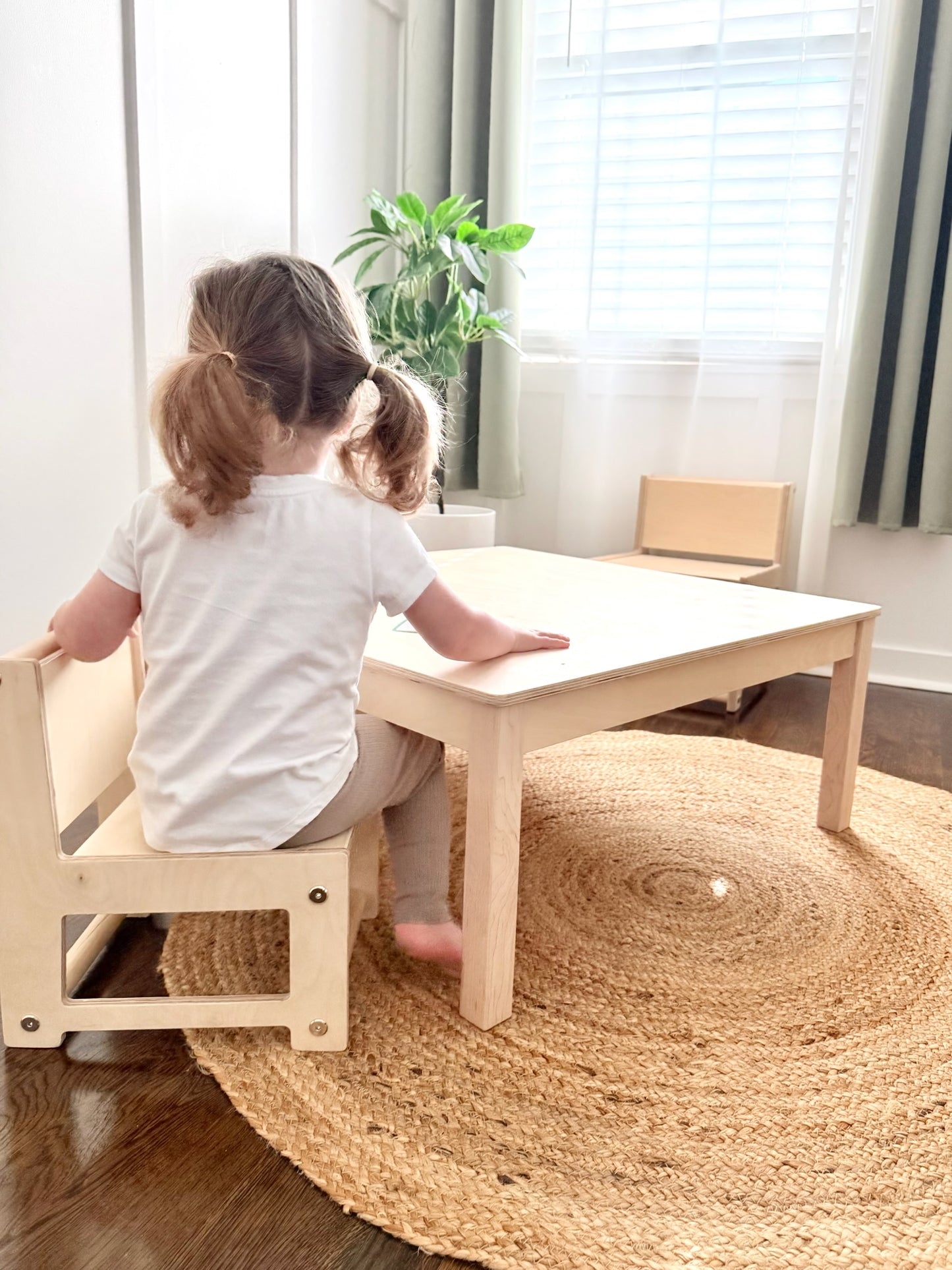 A child sitting at a small wooden montessori table in a bright room with a plant in the background.