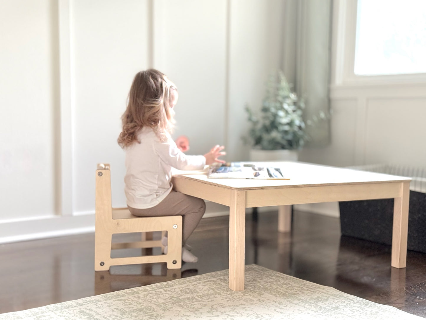 Child sitting at a small wooden Montessori table in a bright room with a plant in the background