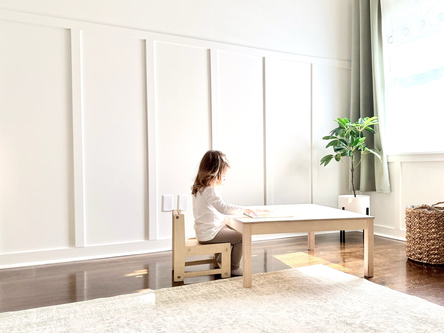 Child sitting at a small wooden table in a bright room with white walls and a plant.