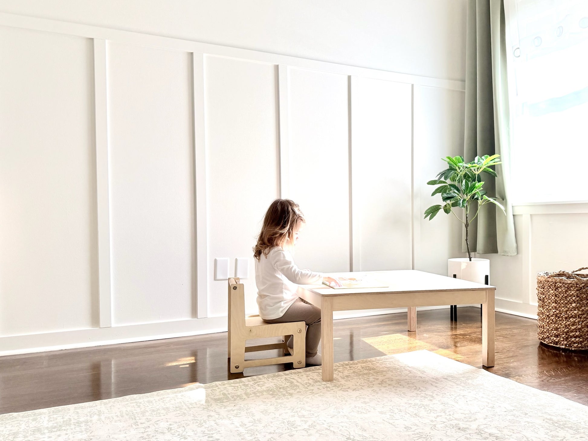 Child sitting at a small wooden table in a bright room with white walls and a plant.